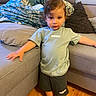 child, barefoot, adidas, tshirt, shorts, couch, pillow, living_room, floor, wooden_floor, person, indoor, furniture, young_child, standing, casual_clothing, home, relaxation, curly_hair, comfort