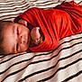 Kai Eli joined the competition — help win amazing prizes! baby, newborn, infant, swaddle, red_swaddle, bracelet, wristband, blanket, striped_blanket, face, eyes, hair, hand, fist, portrait, cozy, lying_down, indoors, soft_texture, cute