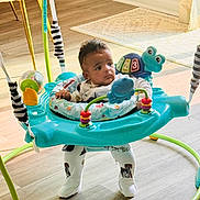 Kai Eli is registered to the contest to win money with this photo: baby, infant, child, jumper, exersaucer, toy, frog_toy, playtime, indoor, hardwood_floor, sunlight, rug, onesie, socks, legs, seat, curtain, wooden_floor, colorful, portrait