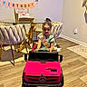 child, birthday, birthday_banner, birthday_cake, toy_car, pink_car, mercedes, crown, sash, girl, sitting, indoors, dining_room, chairs, glass_table, wood_floor, smiling, portrait, party_decorations, celebration