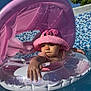 baby, pink_hat, inflatable_float, pool, water, sunlight, child, summer, outdoor, blue_sky, mosaic_tiles, green_tree, cute, reaching_hand, swimming, fun, vacation, sun_hat, infant, playful