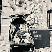 Leewene a rejoint le concours — aidez-le/la à gagner de superbes lots ! baby, stroller, stuffed_toy, statue, black_and_white, outdoor, child, smiling, building, stone_wall, window, sidewalk, person, seat, toy, urban, public_space, happy, infant, portrait