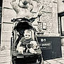 baby, stroller, stuffed_toy, statue, black_and_white, outdoor, child, smiling, building, stone_wall, window, sidewalk, person, seat, toy, urban, public_space, happy, infant, portrait
