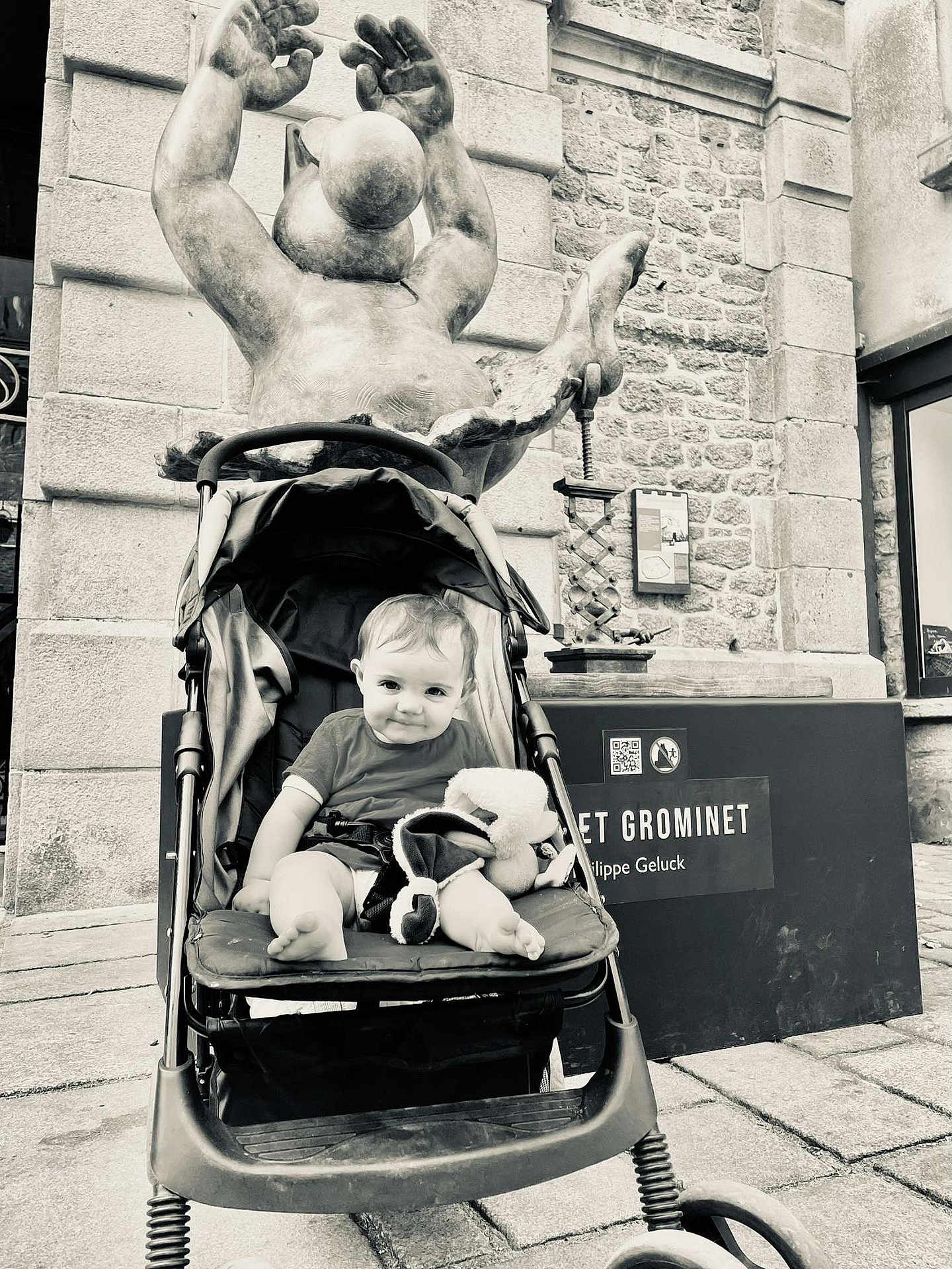 Leewene a rejoint le concours — aidez-le/la à gagner de superbes lots ! baby, stroller, stuffed_toy, statue, black_and_white, outdoor, child, smiling, building, stone_wall, window, sidewalk, person, seat, toy, urban, public_space, happy, infant, portrait