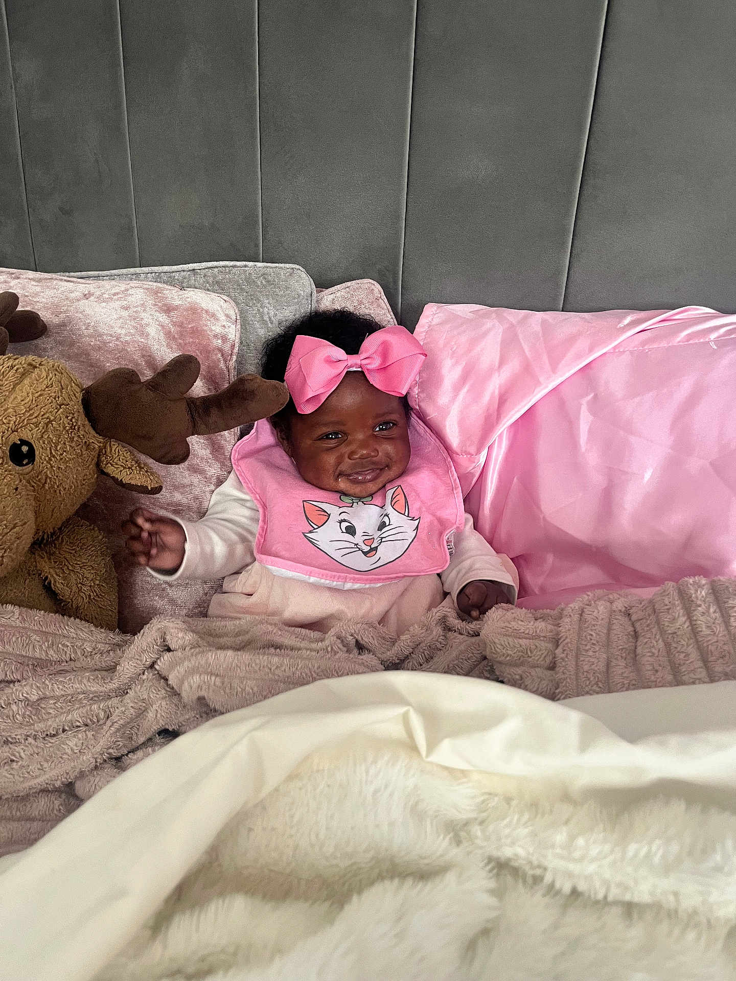 Psalms-Marie is registered to the contest to win money with this photo: baby, child, pink_bow, bib, cartoon_cat, plush_toy, moose, blanket, pillow, bed, headboard, smile, indoor, cozy, soft_texture, happy, clothing, infant, person, portrait