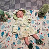 baby, child, dinosaur, robe, blanket, plush_toy, footwear, smile, pattern, bedspread, cute, indoor, soft_toy, infant, comfort, sleepwear, colorful, happy, lying_down, playful
