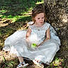 child, girl, white_dress, apple, tree, grass, outdoor, nature, sitting, sunlight, shadows, sandals, young, portrait, casual, daylight, leafy, ground, thoughtful, calm