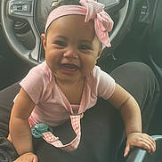 Aviana is registered to the contest to win money with this photo: baby, child, smiling, headband, pink, bow, car_interior, steering_wheel, person, seat, clothing, happy, infant, sitting, lap, cute, young, portrait, close_up, hands