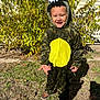child, dinosaur_costume, smiling, outdoor, grass, bushes, sunlight, yellow, green, hood, costume, happy, person, footwear, house, wall, nature, daylight, playful, portrait