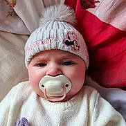 Isla-May is registered to the contest to win money with this photo: baby, bed, beige_blanket, blanket, bow, child, close_up, cute, expression, face, indoors, infant, knitted_hat, minnie_mouse_patch, pacifier, pom_pom, portrait, red_blanket, sitting, white_sweater