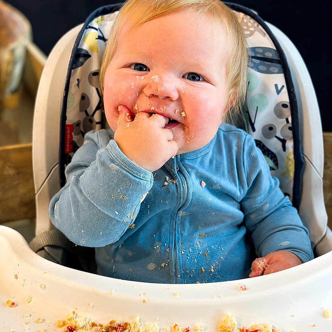 Parker joined the competition — help win amazing prizes! baby, child, high_chair, messy, crumbs, blue_onesie, snack_time, face, hand, smile, happy, indoor, person, cute, infant, food, table, seat, blond_hair, cheeks