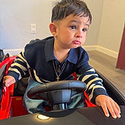 Thiago G is registered to the contest to win money with this photo: child, toddler, boy, portrait, sitting, toy_car, steering_wheel, indoor, smiling, serious_expression, short_hair, sweater, striped_sleeves, necklace, cross_pendant, red_vehicle, hands, face, home, photography