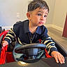 child, toddler, boy, portrait, sitting, toy_car, steering_wheel, indoor, smiling, serious_expression, short_hair, sweater, striped_sleeves, necklace, cross_pendant, red_vehicle, hands, face, home, photography