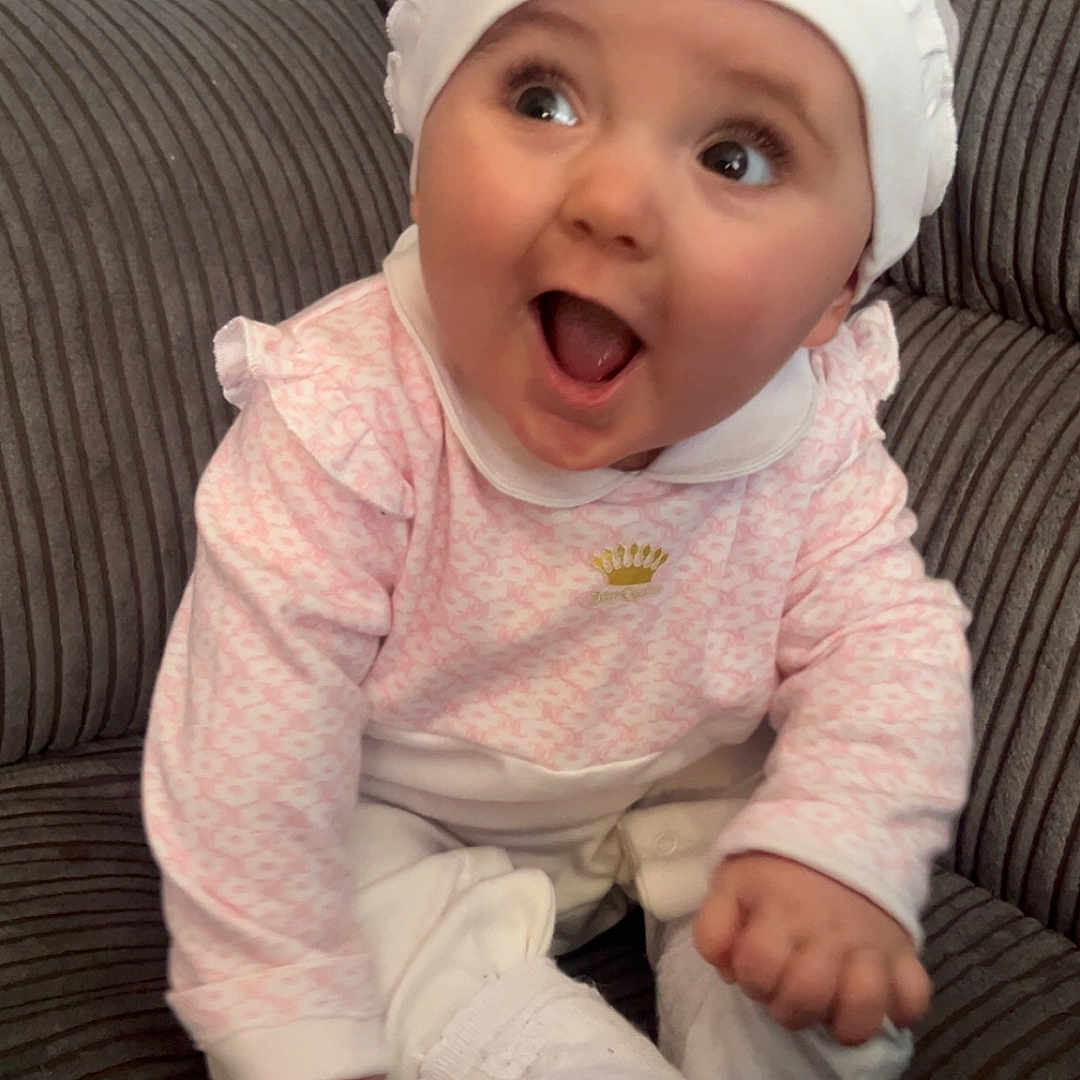 Ivy is registered to the contest to win money with this photo: baby, bodypart, bonnet, cap, clothing, couch, face, finger, furniture, hand, happy, hat, head, mouth, newborn, pants, person, photography, portrait, sitting