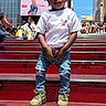 child, urban, city, steps, red, casual_clothing, jeans, tshirt, sneakers, people, buildings, advertisement, daytime, sky, outdoor, portrait, standing, crowd, street, casual