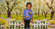Keith is registered to the contest to win money with this photo: child, smiling, outdoor, flowers, fence, grass, orchard, trees, sunlight, spring, nature, plants, happy, person, blue_shirt, black_pants, sneakers, colorful, daytime, greenery
