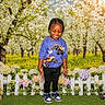 child, smiling, outdoor, flowers, fence, grass, orchard, trees, sunlight, spring, nature, plants, happy, person, blue_shirt, black_pants, sneakers, colorful, daytime, greenery