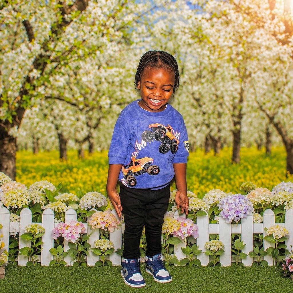 Keith is registered to the contest to win money with this photo: child, smiling, outdoor, flowers, fence, grass, orchard, trees, sunlight, spring, nature, plants, happy, person, blue_shirt, black_pants, sneakers, colorful, daytime, greenery