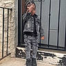 Keith is registered to the contest to win money with this photo: child, denim_jacket, denim_pants, black_shoes, standing, outdoor, concrete, metal_gate, textured_wall, casual_clothing, young_child, fashion, urban, portrait, person, curly_hair, black_top, sidewalk, playful, hand_on_face