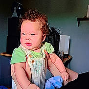 Styles joined the competition — help win amazing prizes! baby, child, curly_hair, green_shirt, overalls, blanket, indoor, person, face, arms, holding, adult_arm, furniture, shelf, electronics, dim_light, portrait, cute, infant, expression
