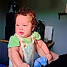 baby, child, curly_hair, green_shirt, overalls, blanket, indoor, person, face, arms, holding, adult_arm, furniture, shelf, electronics, dim_light, portrait, cute, infant, expression