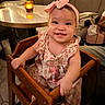 baby, child, smiling, happy, portrait, indoors, restaurant, high_chair, pink_bow, headband, floral_dress, sitting, wooden_furniture, table, candlelight, dinner, background, infant, toothy_smile, family_dining
