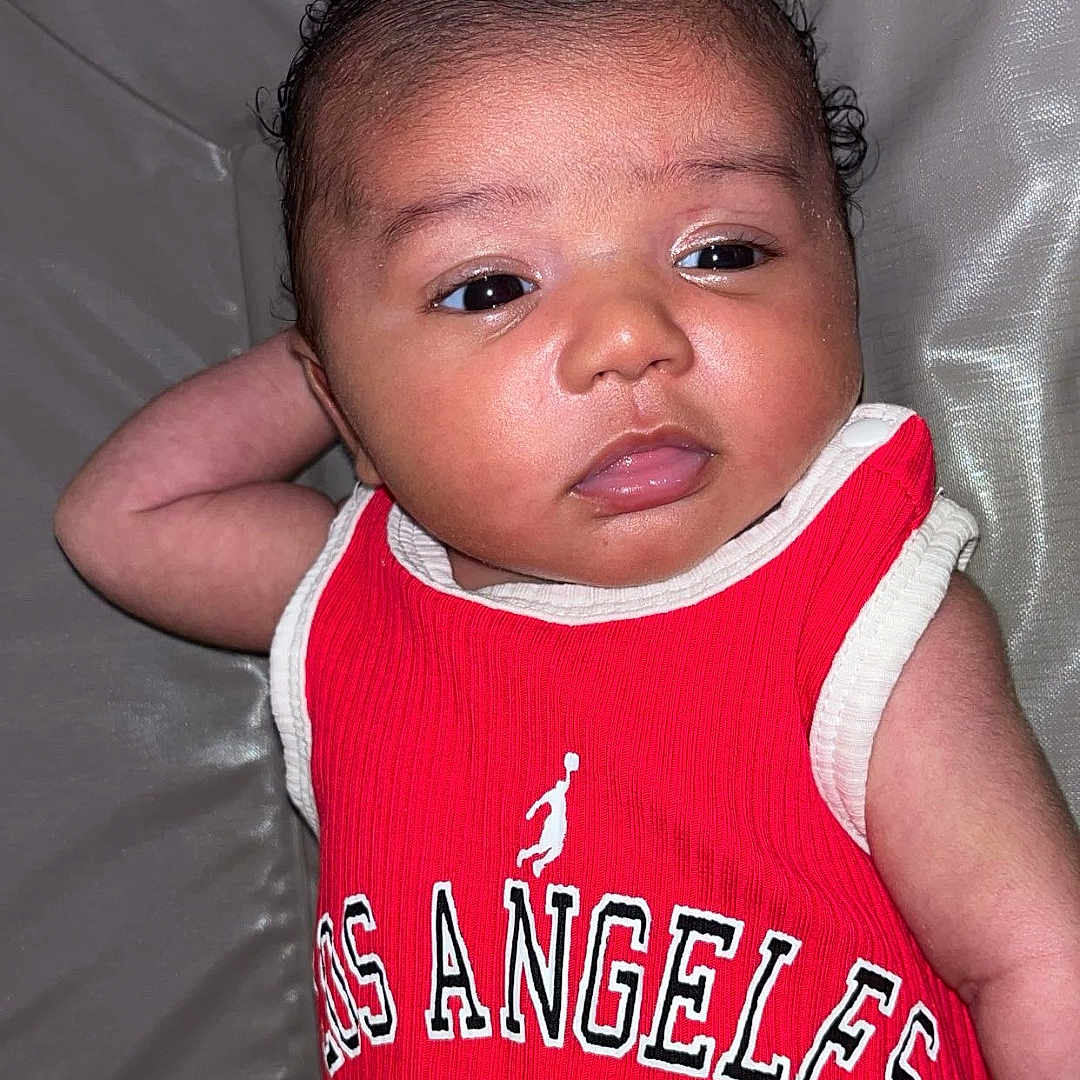 Djeneba a rejoint le concours — aidez-le/la à gagner de superbes lots ! arm, baby, casual, child, clothing, couch, curly_hair, cute, fabric, face, indoors, infant, los_angeles, number_22, person, portrait, red_jersey, relaxed_pose, skin, young