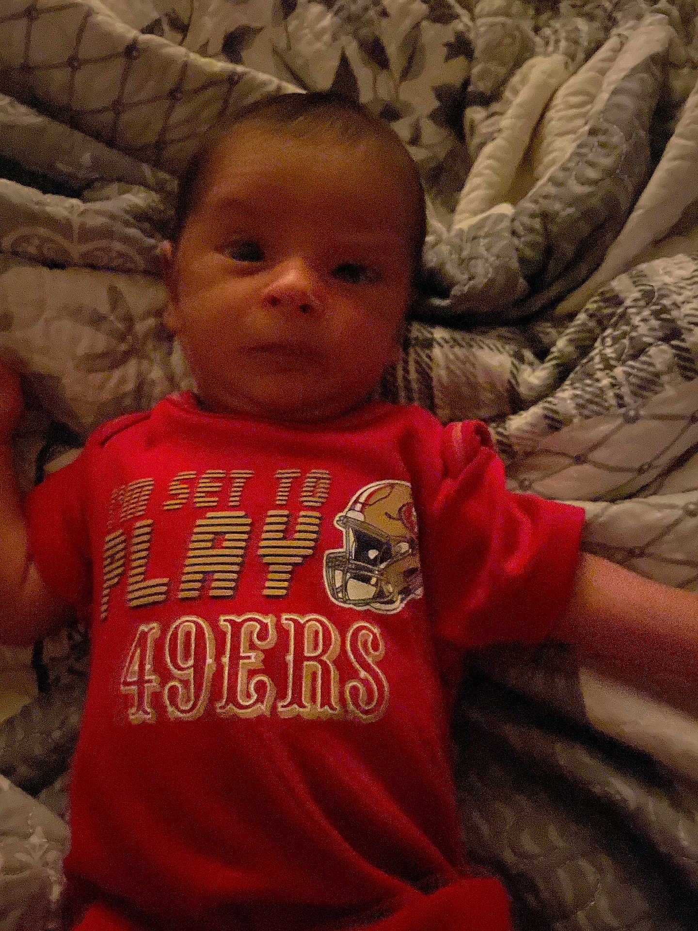 Daemon King Vela is registered to the contest to win money with this photo: baby, infant, child, red_shirt, 49ers, blanket, patterned_blanket, lying_down, indoors, face, head, arm, clothing, fabric, person, portrait, baby_boy, casual, soft_lighting, relaxed