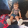 toddler, child, person, sitting, couch, blanket, clothing, tshirt, denim_shorts, sneakers, hair, pigtails, face, blue_eyes, remote_control, indoor, carpet, pattern, cute, smile