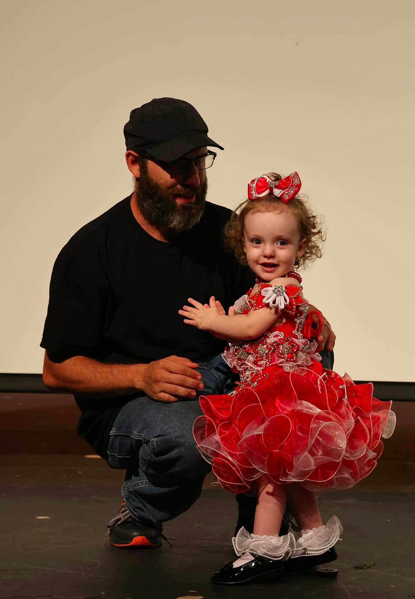 Madilynn joined the competition — help win amazing prizes! child, toddler, man, beard, cap, glasses, red_dress, bow, stage, smile, curly_hair, black_shirt, jeans, black_shoes, white_socks, dance, performance, affection, family, indoor