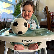 Neo is registered to the contest to win money with this photo: baby, child, infant, high_chair, soccer_ball, football, smiling, portrait, indoor, home, kitchen, window, plants, baby_clothes, bare_feet, happy, seated, playtime, table, soft_lighting