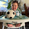 baby, child, infant, high_chair, soccer_ball, football, smiling, portrait, indoor, home, kitchen, window, plants, baby_clothes, bare_feet, happy, seated, playtime, table, soft_lighting