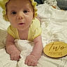 baby, infant, yellow_bonnet, yellow_clothing, white_blanket, wooden_plaque, milestone, two_months, lying_down, bedroom, soft_lighting, curious_expression, close_up, person, child, cute, indoors, portrait, newborn, hands