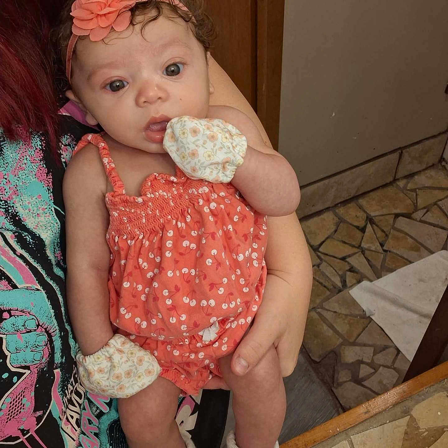 Gabriella joined the competition — help win amazing prizes! baby, infant, child, portrait, sitting, held, face, wide_eyed, looking_at_camera, flower_headband, coral_dress, floral_clothing, mittens, socks, adult, indoor, mosaic_floor, wooden_door, home, cute