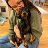 Razmotte a rejoint le concours — aidez-le/la à gagner de superbes lots ! indoor, person, dog, dachshund, glasses, green_sweater, jeans, brown_floor, hugging, pet, cute, cozy, home, sitting, affection, animal, portrait, domestic, companion, casual