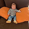 Marius a rejoint le concours — aidez-le/la à gagner de superbes lots ! baby, child, striped_shirt, jeans, orange_pillows, brown_couch, indoor, seated, footwear, casual_clothing, infant, person, cute, home, relaxed, furniture, cozy, young, portrait, comfortable