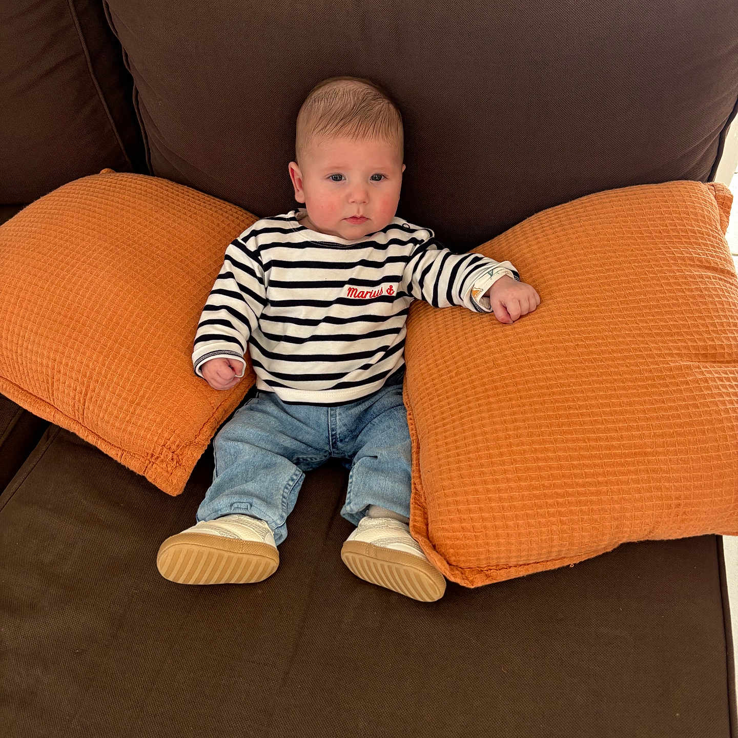 Marius a rejoint le concours — aidez-le/la à gagner de superbes lots ! baby, child, striped_shirt, jeans, orange_pillows, brown_couch, indoor, seated, footwear, casual_clothing, infant, person, cute, home, relaxed, furniture, cozy, young, portrait, comfortable