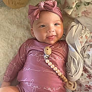 Adalyn is registered to the contest to win money with this photo: baby, infant, smiling, headband, pink_clothing, blanket, wooden_name_plaque, lying_down, cute, indoors, soft_texture, pacifier_clip, child, person, portrait, happy, cozy, fabric, newborn, warm