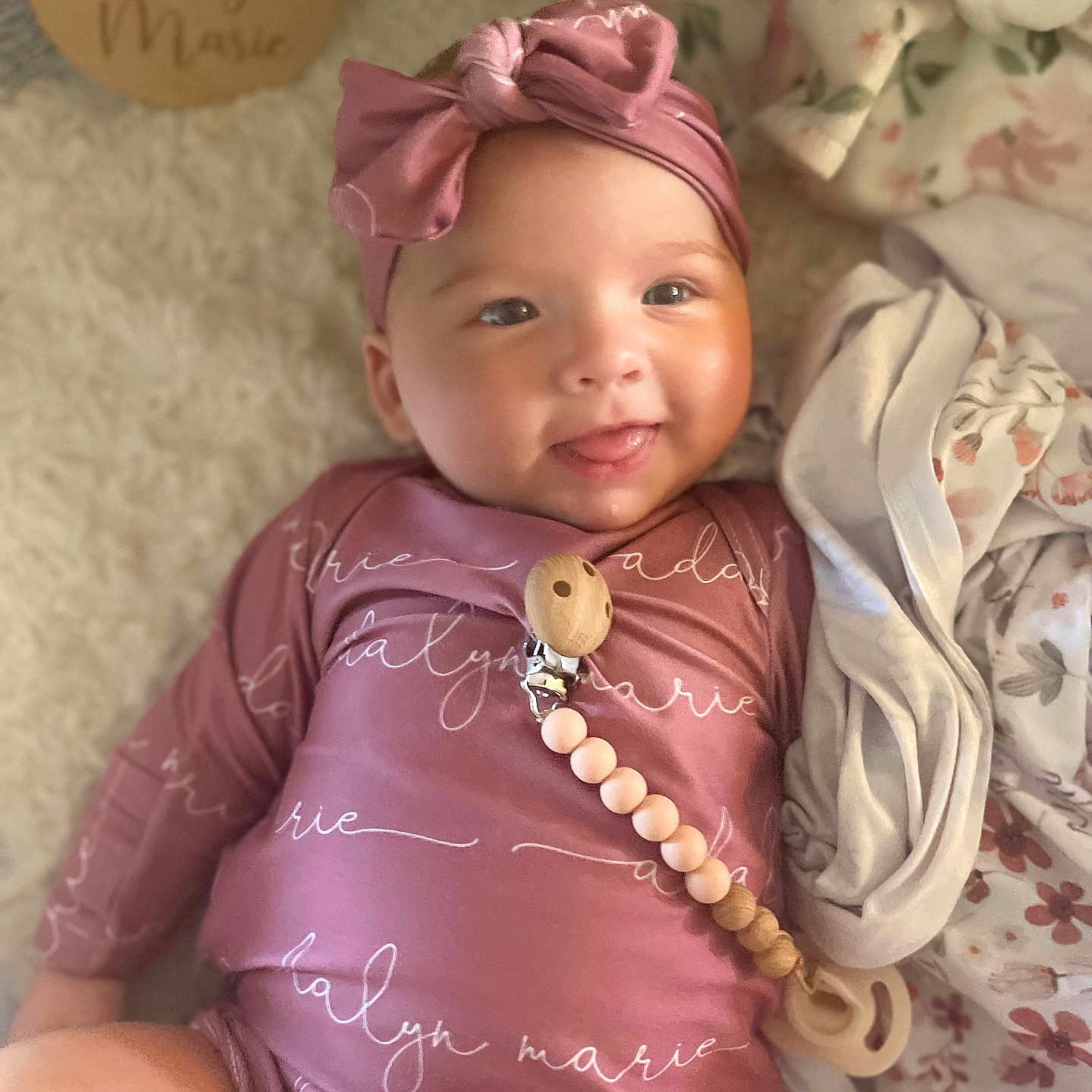 Adalyn is registered to the contest to win money with this photo: baby, blanket, child, cozy, cute, fabric, happy, headband, indoors, infant, lying_down, newborn, pacifier_clip, person, pink_clothing, portrait, smiling, soft_texture, warm, wooden_name_plaque