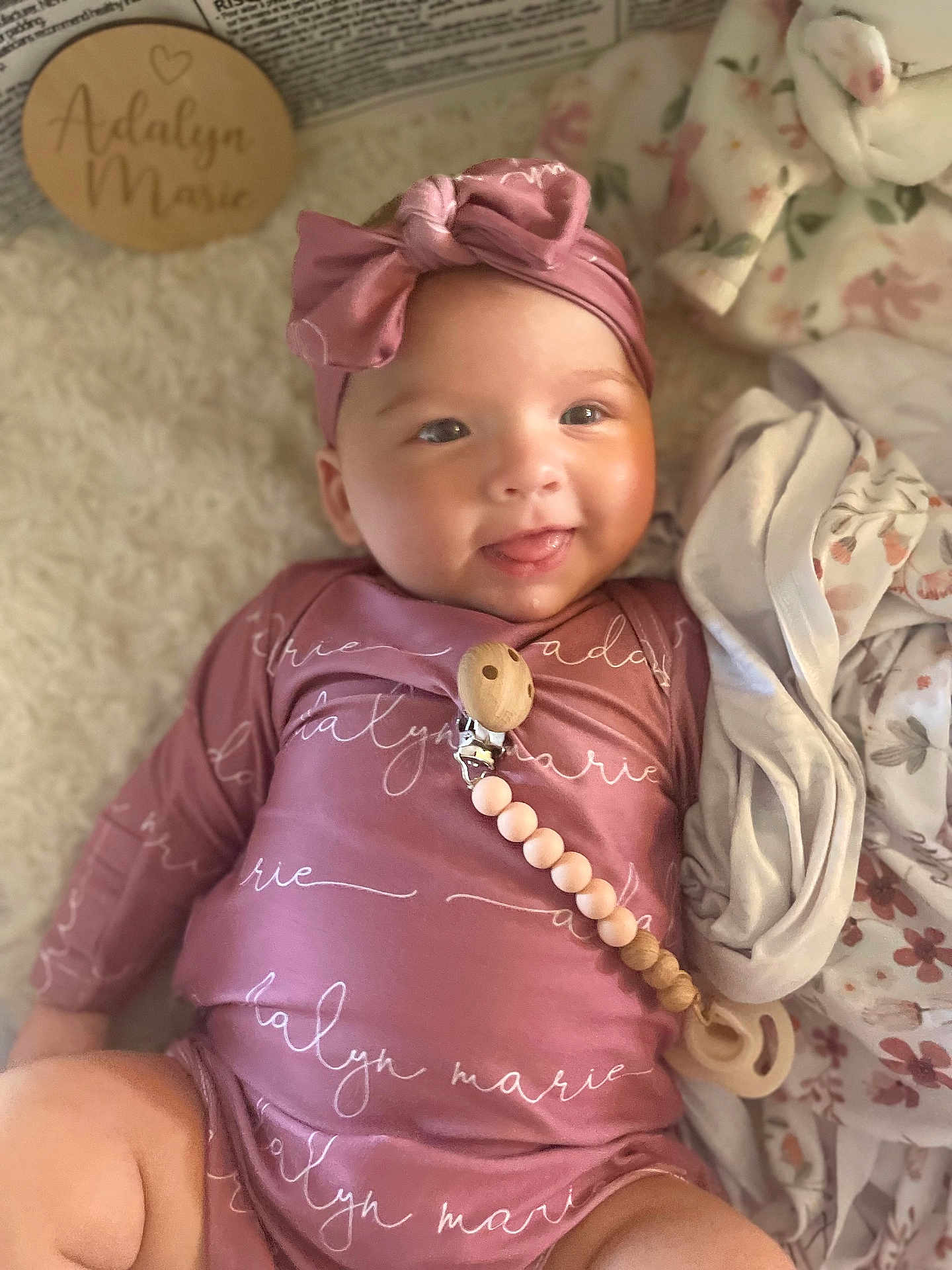 Adalyn is registered to the contest to win money with this photo: baby, infant, smiling, headband, pink_clothing, blanket, wooden_name_plaque, lying_down, cute, indoors, soft_texture, pacifier_clip, child, person, portrait, happy, cozy, fabric, newborn, warm