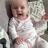 Romi joined the competition — help win amazing prizes! baby, infant, smiling, onesie, cartoon_print, couch, pillow, blanket, indoor, child, happy, cute, hand, face, person, seated, cozy, living_room, apparel, portrait