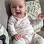 baby, infant, smiling, onesie, cartoon_print, couch, pillow, blanket, indoor, child, happy, cute, hand, face, person, seated, cozy, living_room, apparel, portrait