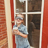 Sailor is registered to the contest to win money with this photo: child, girl, denim_dress, sunglasses, cup, door, window, reflection, brick_wall, smiling, outdoor, standing, building, casual, fashion, young, happy, person, daylight, urban
