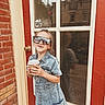 child, girl, denim_dress, sunglasses, cup, door, window, reflection, brick_wall, smiling, outdoor, standing, building, casual, fashion, young, happy, person, daylight, urban