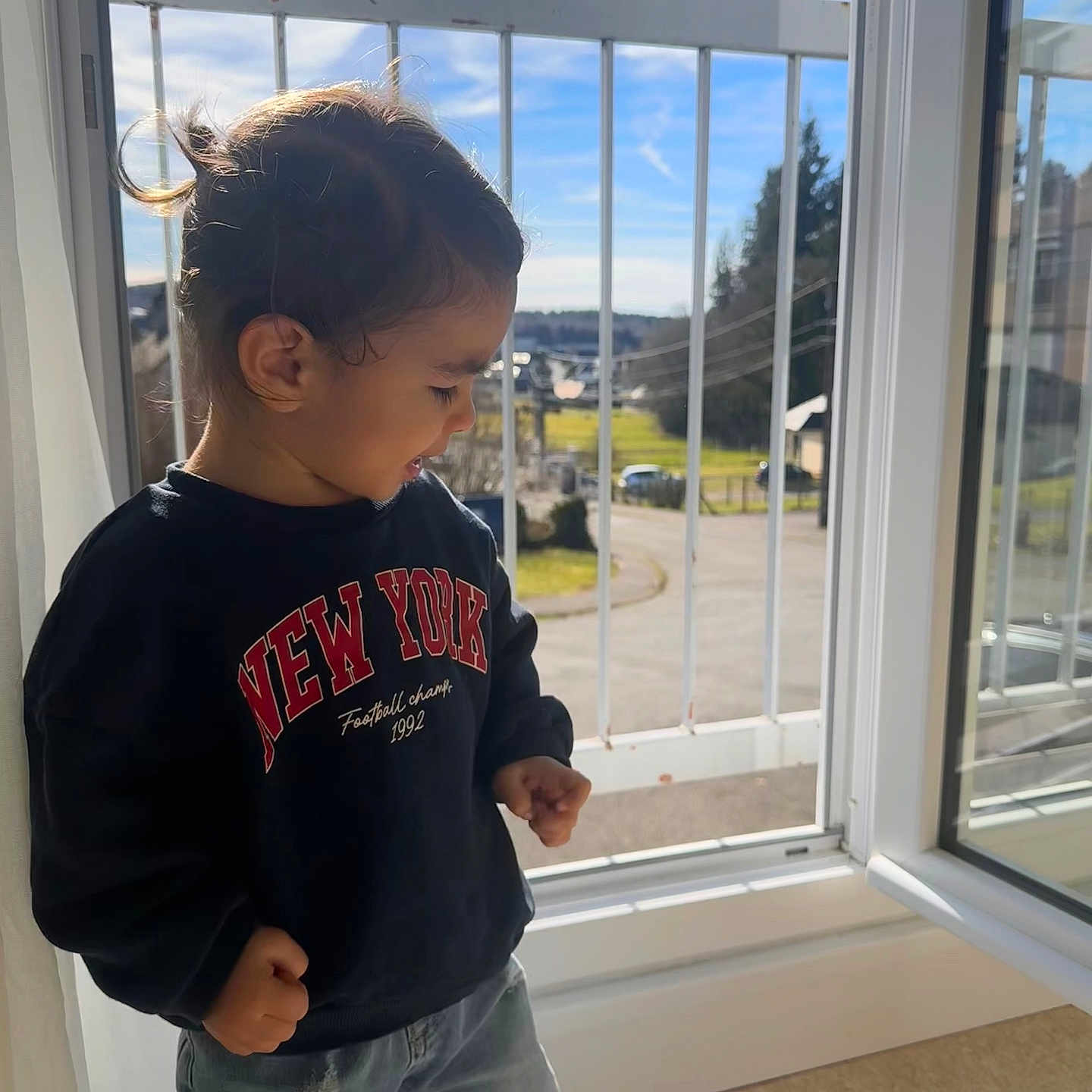 Batuhan a rejoint le concours — aidez-le/la à gagner de superbes lots ! child, toddler, sweatshirt, jeans, window, open_window, daylight, sunlight, indoor, balcony, railing, smiling, happy, casual_clothing, portrait, young_child, curly_hair, side_view, playing, home