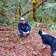 Lilbit is registered to the contest to win money with this photo: man, dog, forest, moss, leaves, tree, nature, outdoor, crouching, pet, canine, greenery, fern, fall, woods, animal, person, hat, casual_clothing, leaf_litter