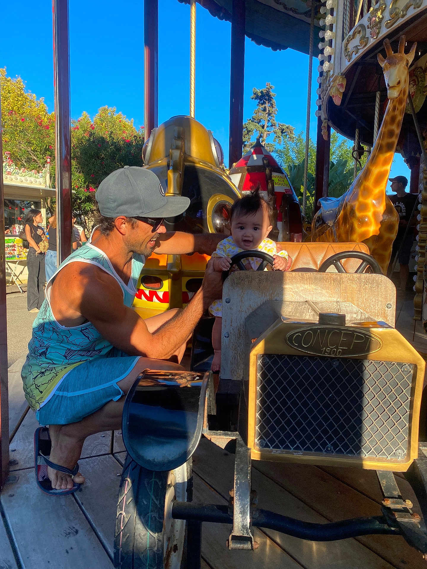 Heipoe Fontaine a rejoint le concours — aidez-le/la à gagner de superbes lots ! child, man, carousel, amusement_park, toy_car, giraffe, steering_wheel, sunlight, outdoor, cap, sunglasses, smile, happy, person, ride, summer, play, fun, family, vacation