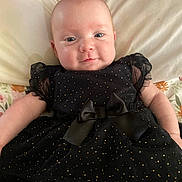Odessa is registered to the contest to win money with this photo: baby, infant, black_dress, bow, bed, pillow, floral_sheet, smile, face, head, cute, child, person, lying_down, indoors, soft_lighting, portrait, newborn, clothing, happy