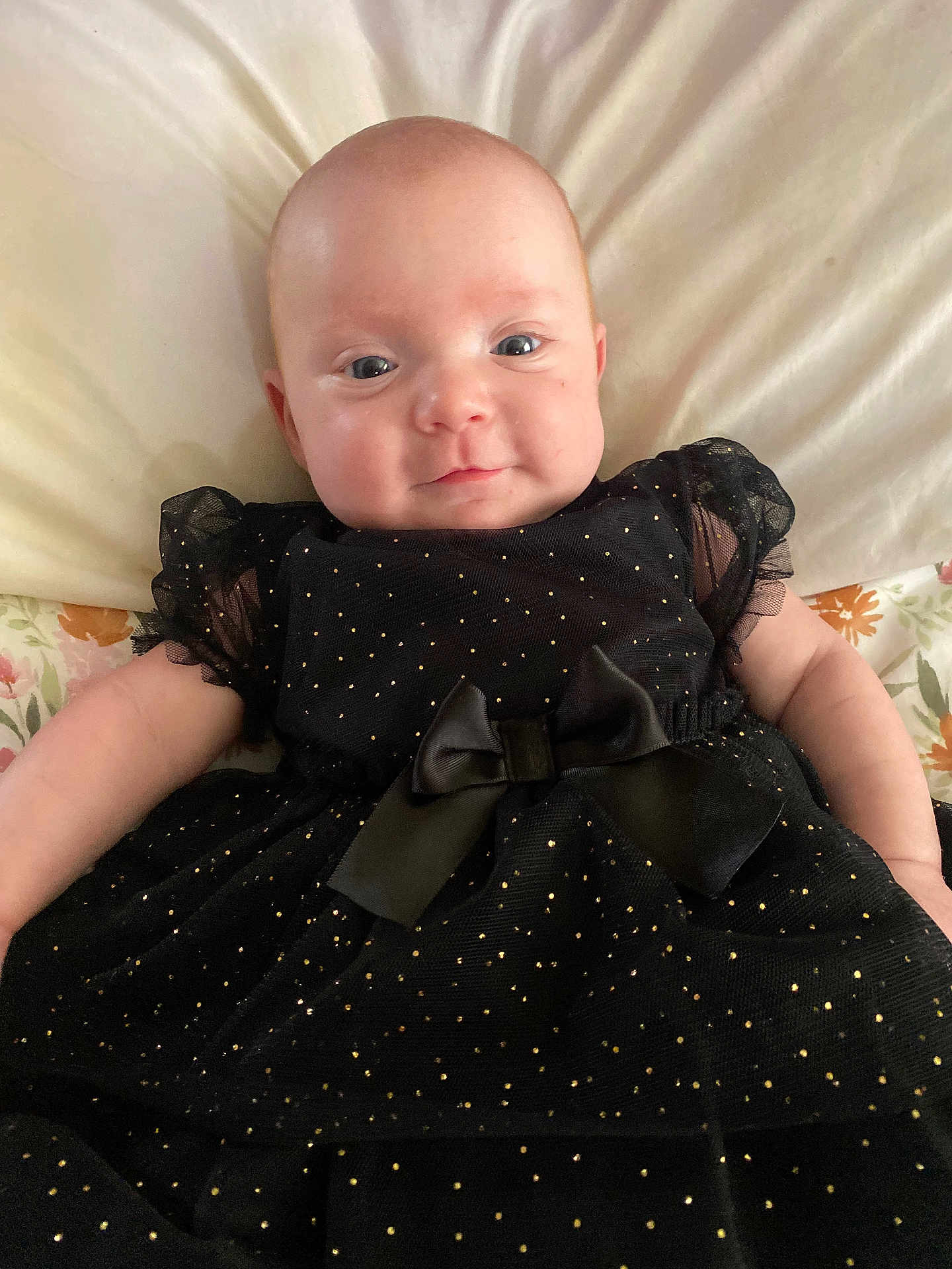 Odessa is registered to the contest to win money with this photo: baby, infant, black_dress, bow, bed, pillow, floral_sheet, smile, face, head, cute, child, person, lying_down, indoors, soft_lighting, portrait, newborn, clothing, happy
