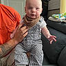 baby, child, person, overalls, bib, tattoo, hand, couch, blanket, window, door, indoor, smile, expression, sitting, clothing, furniture, home, adult_hand, striped_clothing